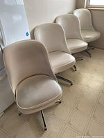 Four cream-colored swivel chairs with upholstered rounded seats and backs arranged in a row on a tiled floor beside a refrigerator.