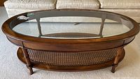 Front view of the oval coffee table showing the wood frame, glass top, and cane shelf underneath.