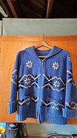 Full front view of blue knit Cowichan sweater with snowflake and geometric patterns, hanging on wooden hanger against wood background.
