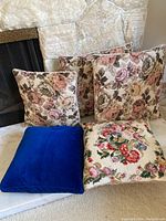 Set of five throw pillows on a fireplace hearth including three floral fabric pillows, one blue velvet pillow, and one multicolor floral tapestry pillow.