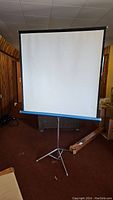 Full view of the white projector screen mounted on the adjustable tripod stand inside a room with wood panel walls.
