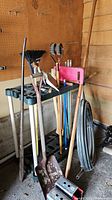 Garden tools such as shovels, broom, rake, and others in a black plastic storage rack, with coiled pipe tubing beside it.