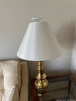 Full view of brass table lamp with white shade beside sofa and on a glass-top wooden side table, showing base and shade details.