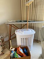 Wooden ironing board with gray cover, wooden carpet rake, white plastic laundry basket with a lid, cardboard box with cleaning cloths and cleaning supplies bottles.