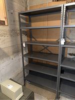 Full view of the metal shelf unit showing six shelves, standing against a basement wall with some cardboard behind.