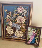 Both framed petit point needlepoint artwork pieces positioned on a carpeted floor, one large floral bouquet and one smaller woman with dog.