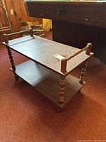Wooden rolling cart shown front left angle on carpeted floor, featuring two shelves and caster wheels.