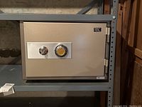 Full view of beige TDL safe on gray metal shelving unit showcasing front with combination dial and key lock.