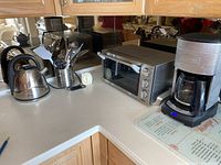 Corner kitchen countertop with Oster kettle, Delonghi toaster oven, and Cuisinart coffee maker placed side by side.