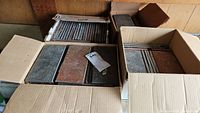 Four open cardboard boxes containing rectangular floor tiles in dark gray and reddish brown colors. Tiles measure approximately 8x4 inches with about 0.5 inch thickness.