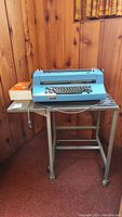 Blue IBM electric typewriter sitting on a metal frame table with wood top and power cord visible.