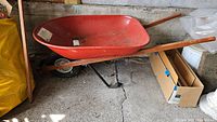 Side view of the red metal tray, wooden handle, and part of the wheelbarrow wheel attached to black metal frame, placed on a garage floor.