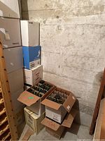 View of multiple stacked cardboard boxes with empty green wine bottles inside, set in a concrete basement next to a wooden wine rack.