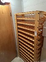 Full view of a tall wooden wine rack in a corner with natural wooden finish, showing several rows of horizontal slots for storing wine bottles.