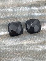 Two faceted cushion-cut black sapphire stones placed side by side on a textured fabric background, showing their faceting and reflective qualities.
