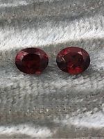 Two oval-shaped garnet gemstones shown from above on textured fabric background, deep red color, faceted surface visible.