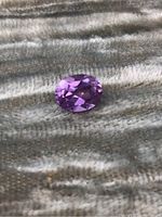 Clear close-up photo of an oval-shaped purple amethyst gemstone on a textured fabric background showing facets and color.