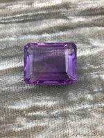 Rectangular emerald-cut amethyst gemstone with rich purple color and visible inclusions, placed on a gray textured surface.