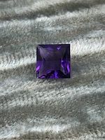 Top view of square-cut amethyst gemstone on textured background, showing rich deep purple color and faceting.