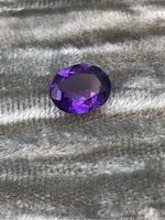 Close-up image of the oval faceted amethyst gemstone on a textured surface showing its deep purple color and faceting.