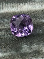 Close-up image of a cushion-cut amethyst gemstone showing its purple color and faceting details on a textured surface.