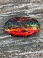 Image of oval shaped, faceted Ammolite gemstone with vibrant iridescent colors on a gray fabric background.