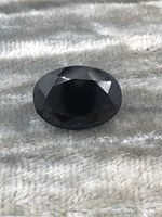 Close-up image of an oval faceted black sapphire gemstone placed on a light textured fabric showing the top view of the gem.
