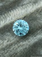 Close-up photo of a round brilliant cut blue moissanite gemstone on a textured fabric background showing facets and color.