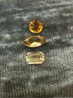 Three loose citrine gemstones placed on fabric, showing different cuts and color shades, with close-up showing facets and clarity.