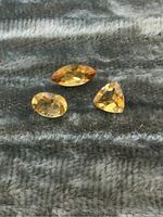 Three loose citrine gemstones displayed on a textured fabric surface: marquise cut, oval cut, and trillion cut stones.