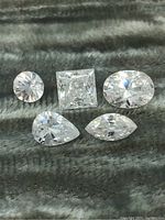 Five loose colourless faceted gemstones of various shapes arranged on a fabric surface. Shapes: round, square, oval, pear, marquise.