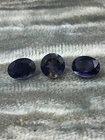 Three oval iolite gemstones laid side by side on a grey fabric background under natural light. Stones show purple-blue hues and faceted cuts.