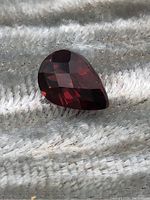 Close-up photo of pear-shaped Mozambique garnet gemstone with dark red faceted surfaces on textured cloth background.