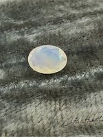 Oval faceted opal gemstone showing milky white translucency with blue iridescence on dark textured fabric background.