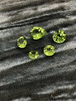 Five loose peridot gemstones including one pear-shaped and four round-cut, displayed on a textured fabric in green tone.