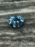 Close-up of oval blue tourmaline gemstone lying on a textured fabric background, showing facets and light reflections.