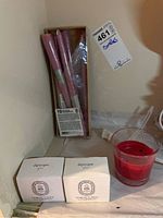Six 10-inch pink taper candles in original box, two unopened Diptyque of Paris brass candlestick holders in white boxes, one red votive candle in a glass holder.