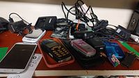 Various cell phones and accessories on a table including flip phones, smartphones, chargers, and cords.