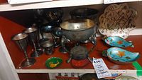 Shelf showing silver-plated wine and champagne glasses, child size cup, coffee pot stands, a small green cloisonne dish, and other metal items