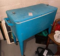 Blue metal party cooler with two lids, embossed 'COLD DRINKS' on the body, metal legs with casters, side handle and bottle opener visible. Shows wear and dirt marks.