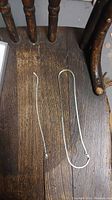 Two thin sterling silver chains, necklace and bracelet, laid flat on a wooden chair seat, showing their shape and condition.