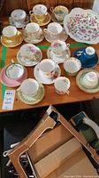 Overview image showing a wooden table with multiple assorted teacups and saucers arranged in groups, featuring various floral and gold rim decorations
