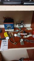 Shelf displaying various watches, watch parts, and clock tins including Fossil branded blue leather wallet clock tin and multiple watches with metal and leather bands. Horse-shaped clock or watch holder included.