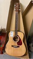 Full view of Yamaha dreadnought acoustic guitar in a cardboard storage box showing body, neck, and headstock with strings and pickguard.