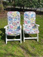 Two white plastic outdoor lounger chairs with floral cushions side by side on grass.