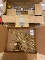 Box containing brass-colored bathroom accessories including towel rings, towel rails, toilet paper holder, and mounting hardware