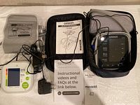 Two digital blood pressure monitors side by side, Omron BP785 in zippered case with gray cuff, Greater Goods with colorful display and cuff, power adapters and manuals visible