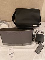 Bose SoundDock speaker, power adapter, remote control, and black carrying case displayed together on a table.