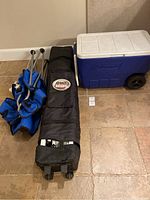 Photo showing folded blue lawn chair, black wheeled bag with Quik Shade logo, and blue Coleman wheeled cooler on tiled floor.