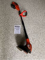Black & Decker orange cordless trimmer/edger with instruction manual on floor showing full tool length and handle.